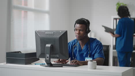 Afroamerikanischer-Männlicher-Arzt-Oder-Krankenschwester-Mit-Headset-Und-Computer,-Der-Im-Krankenhaus-Arbeitet.-Junger-Professioneller-Therapeut,-Arzt,-Der-Den-Kunden-Berät,-Indem-Er-Fernkommunikation-Nutzt-Und-über-Die-Webcam-Spricht